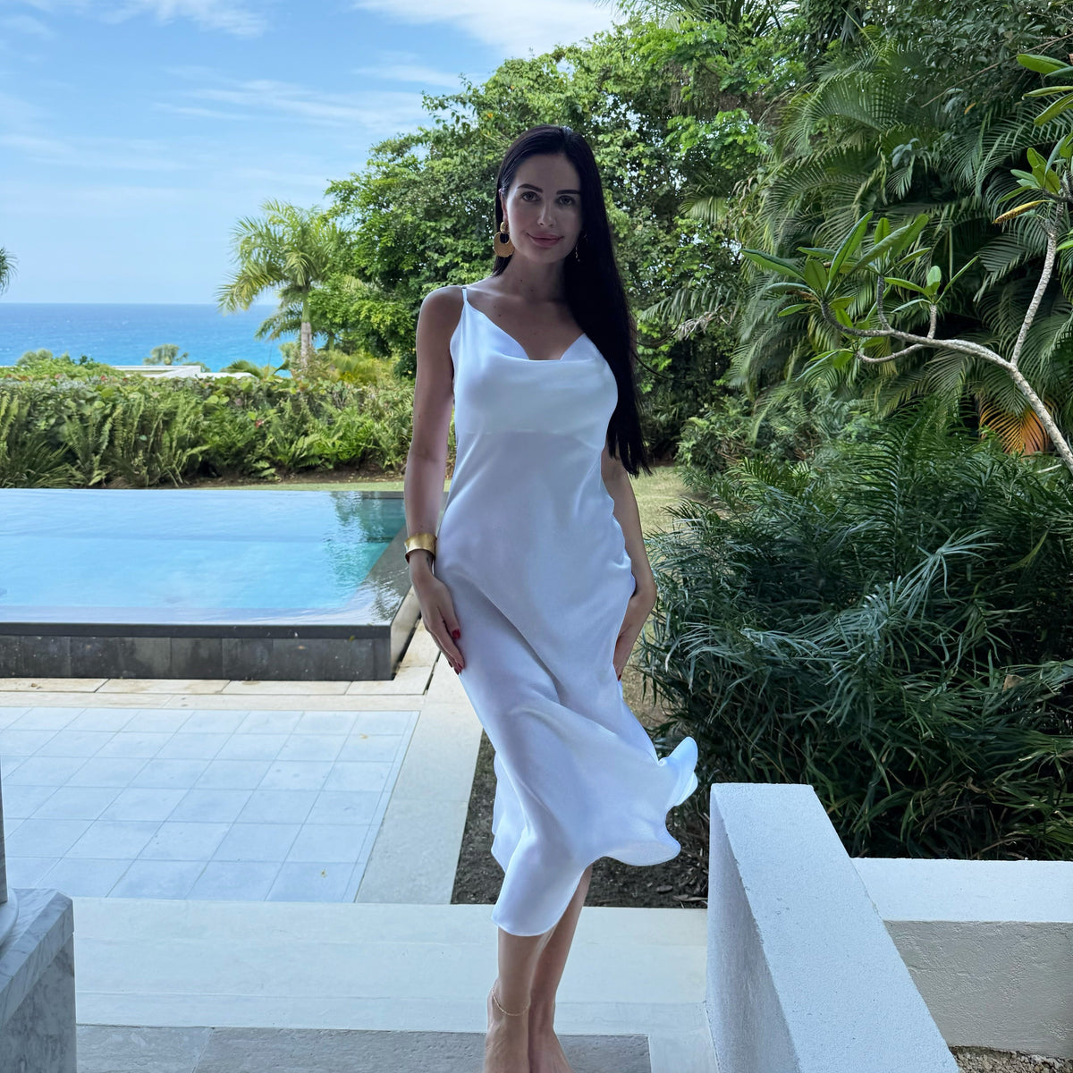 A person wearing a classic white sleeveless silk dress standing near a pool with tropical surroundings.