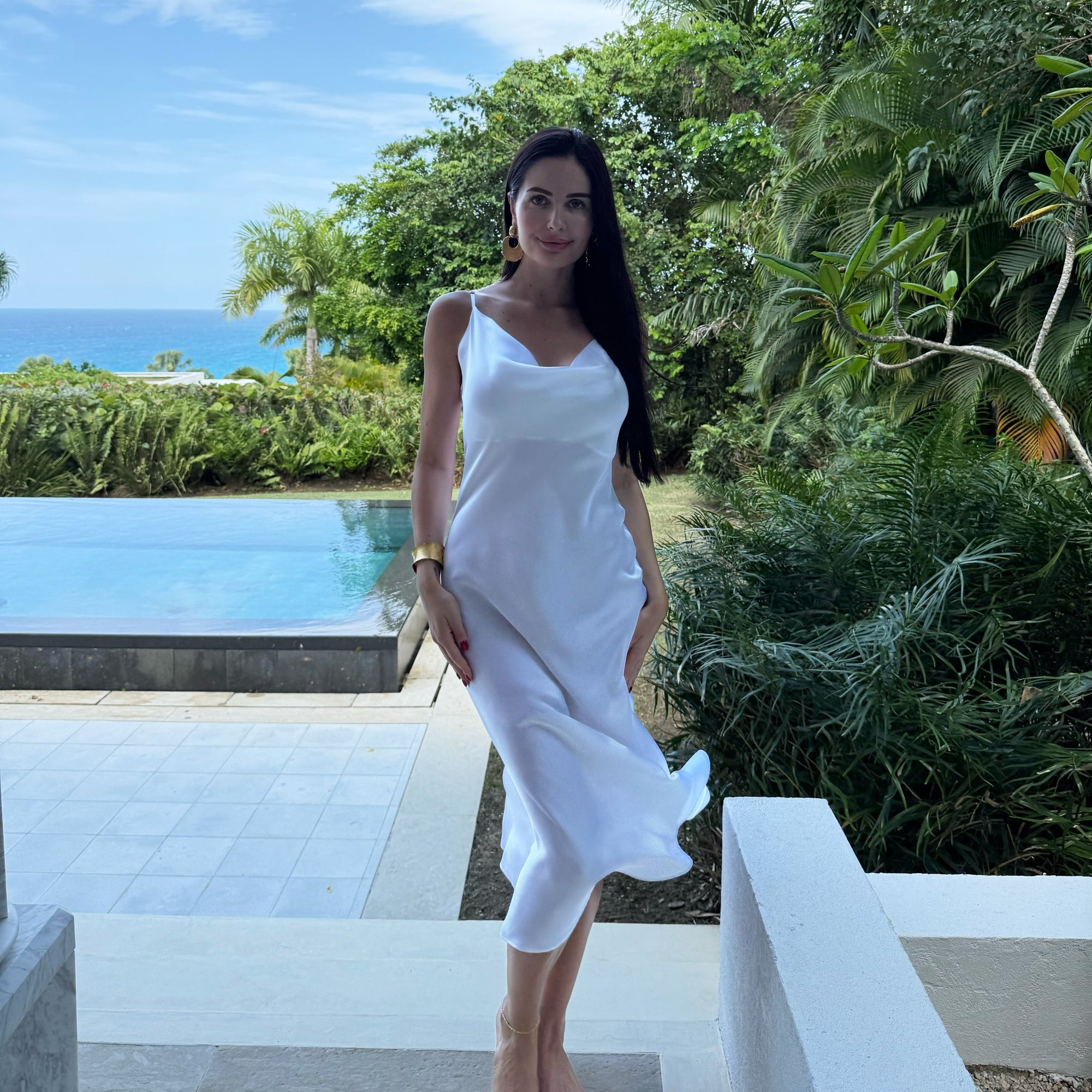A person wearing a classic white sleeveless silk dress standing near a pool with tropical surroundings.